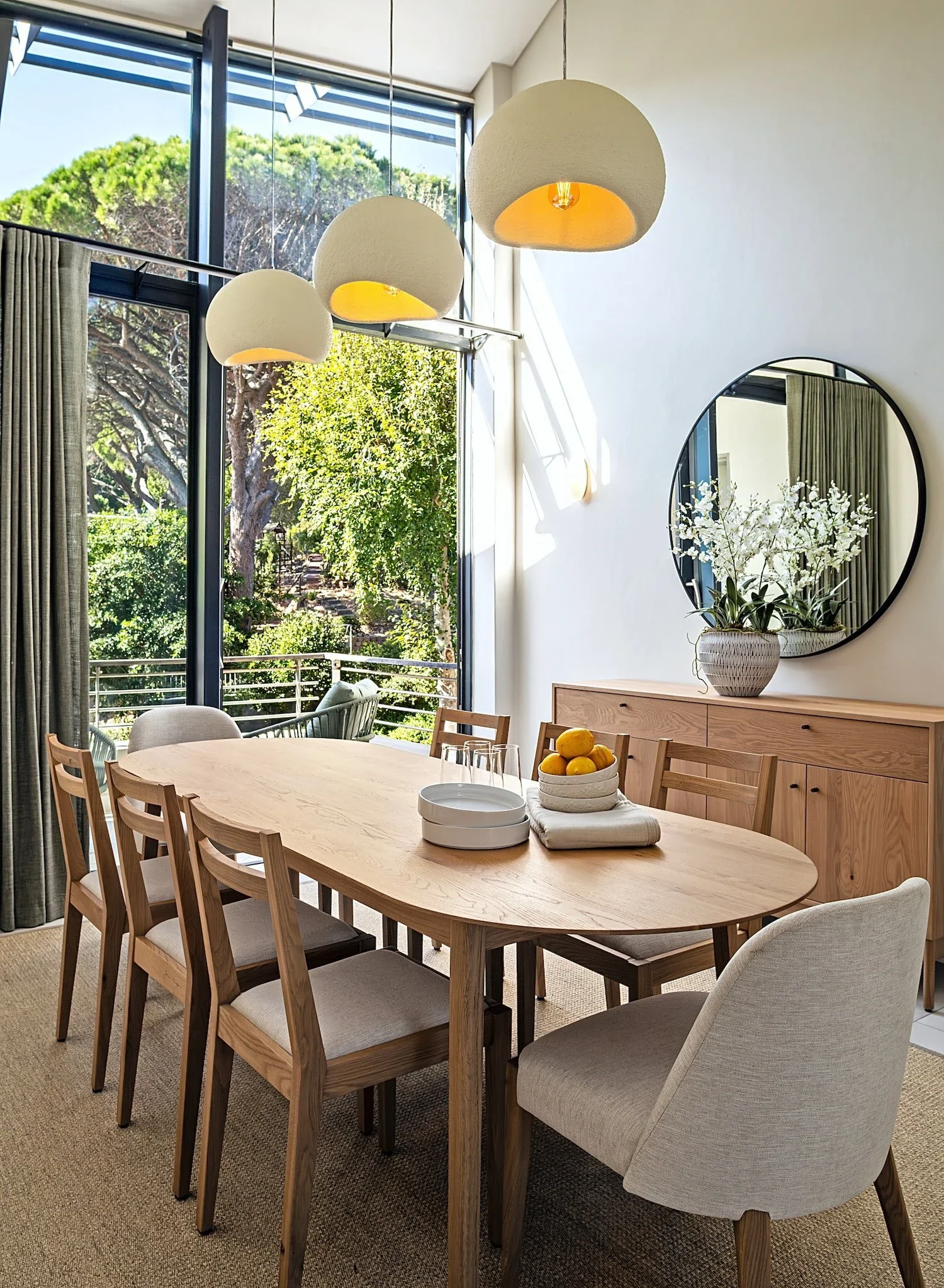 Dining room at The Glen, Camps Bay: an oval oak table and chairs, three cream pendant lights, a round mirror above an oak sideboard and black-framed windows