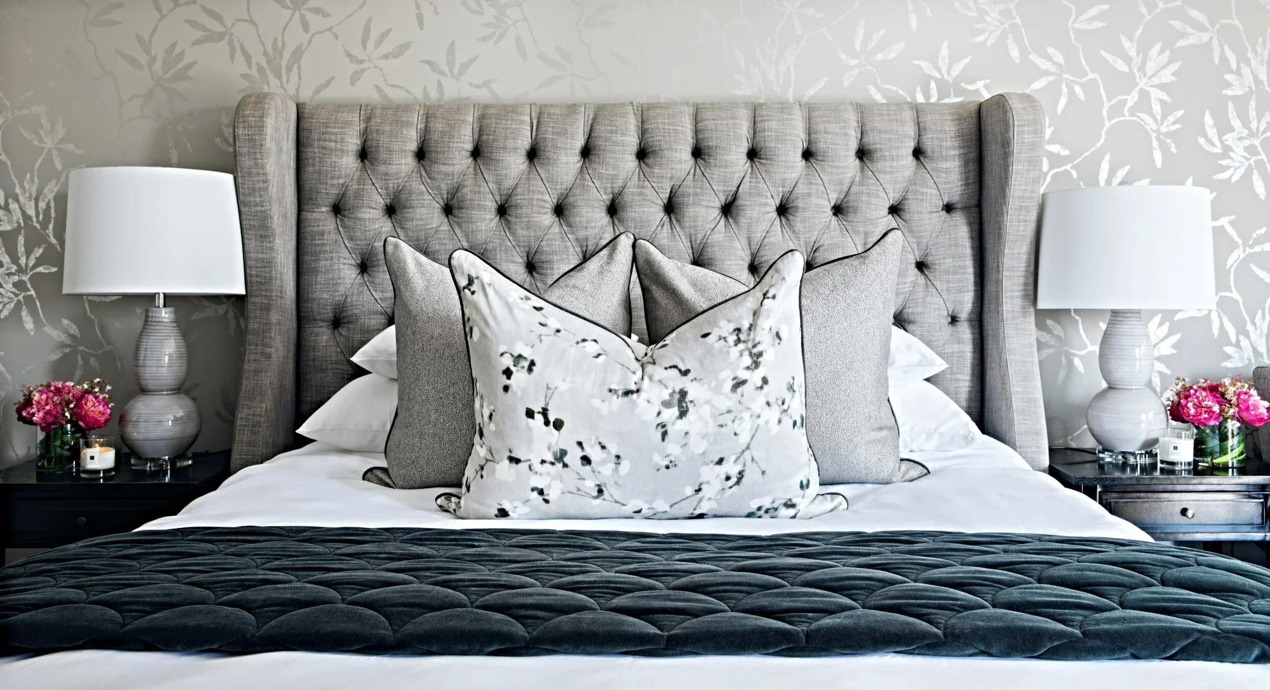 Bedroom with a grey buttoned wingback headboard, botanical cushions, white lamps and silver botanical wallpaper