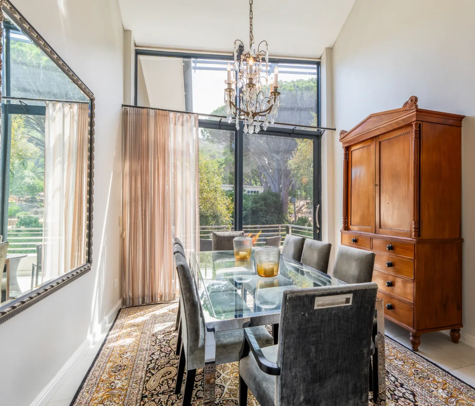 Dining room at The Glen before the work: a crystal chandelier, a glass table with grey velvet chairs, a timber armoire and a patterned rug