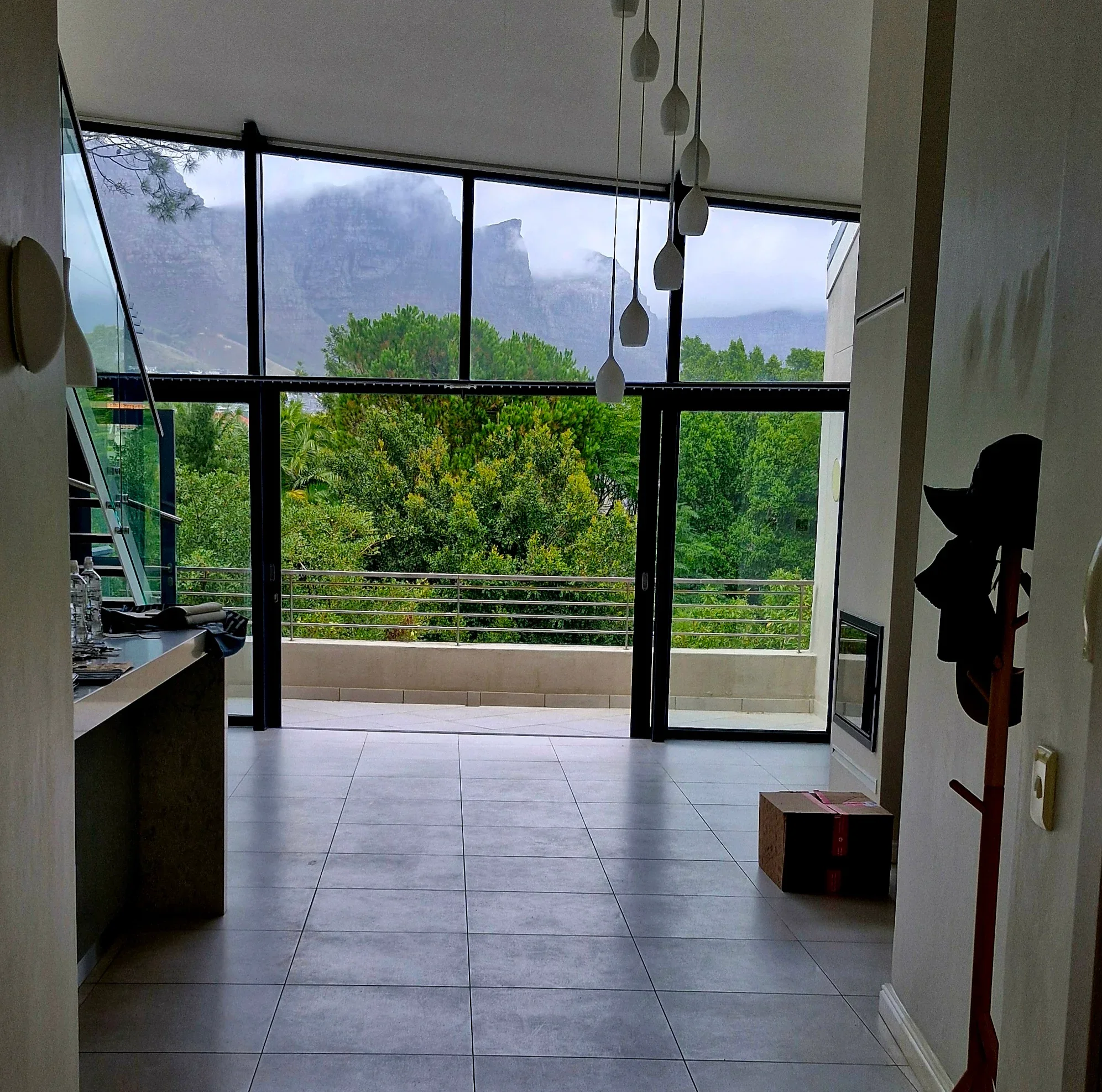 The living room at The Glen before the work: an empty double-height room with tiled floors, glass pendant lights and tall black-framed windows onto the trees and the mountain