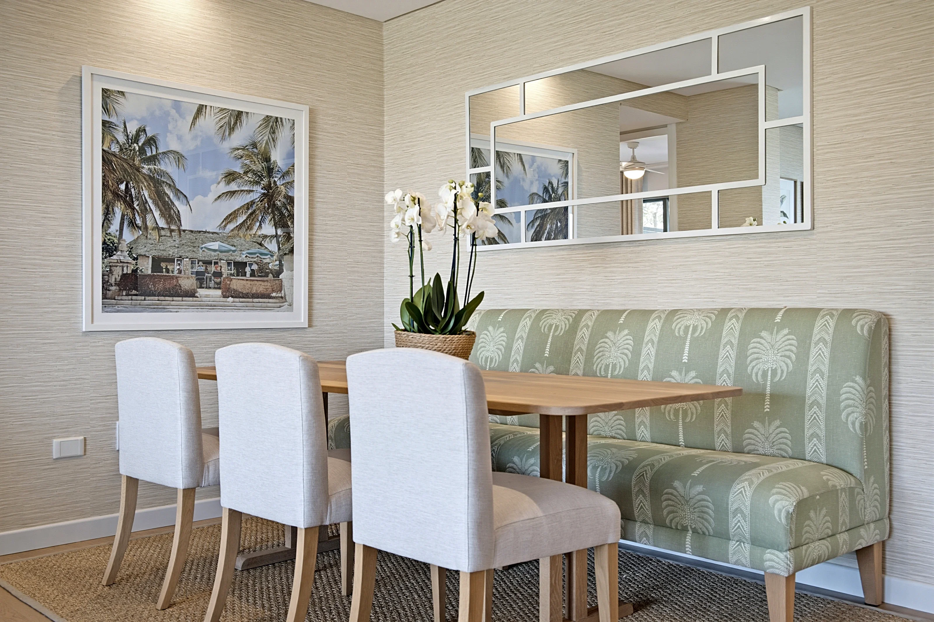 Dining area at Simbithi Eco-Estate: a sage and cream palm-print banquette, an oak pedestal table, white upholstered chairs and a geometric mirror