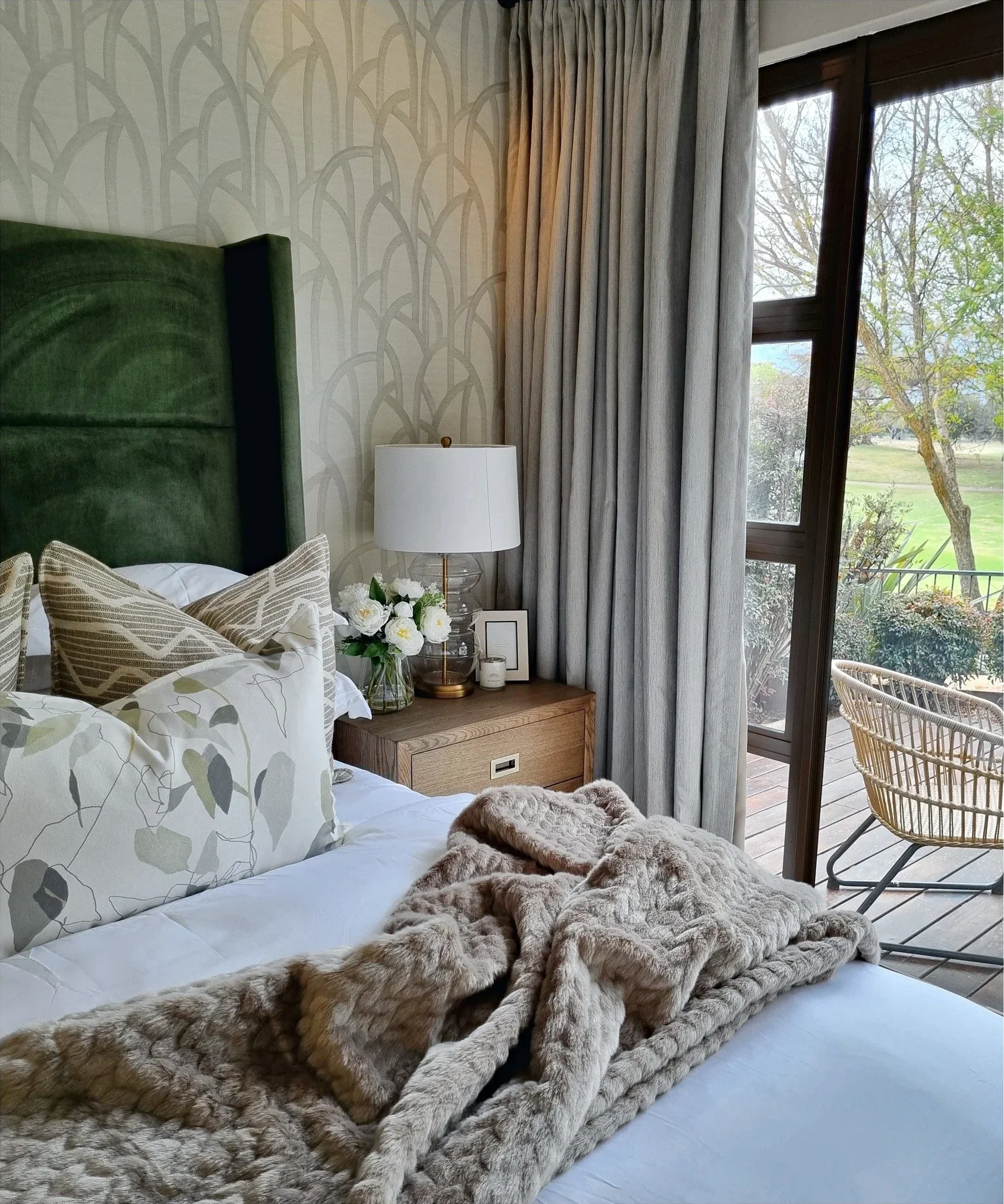 Bedroom with a green velvet headboard, botanical cushions, a knitted throw and a woven chair by the garden door