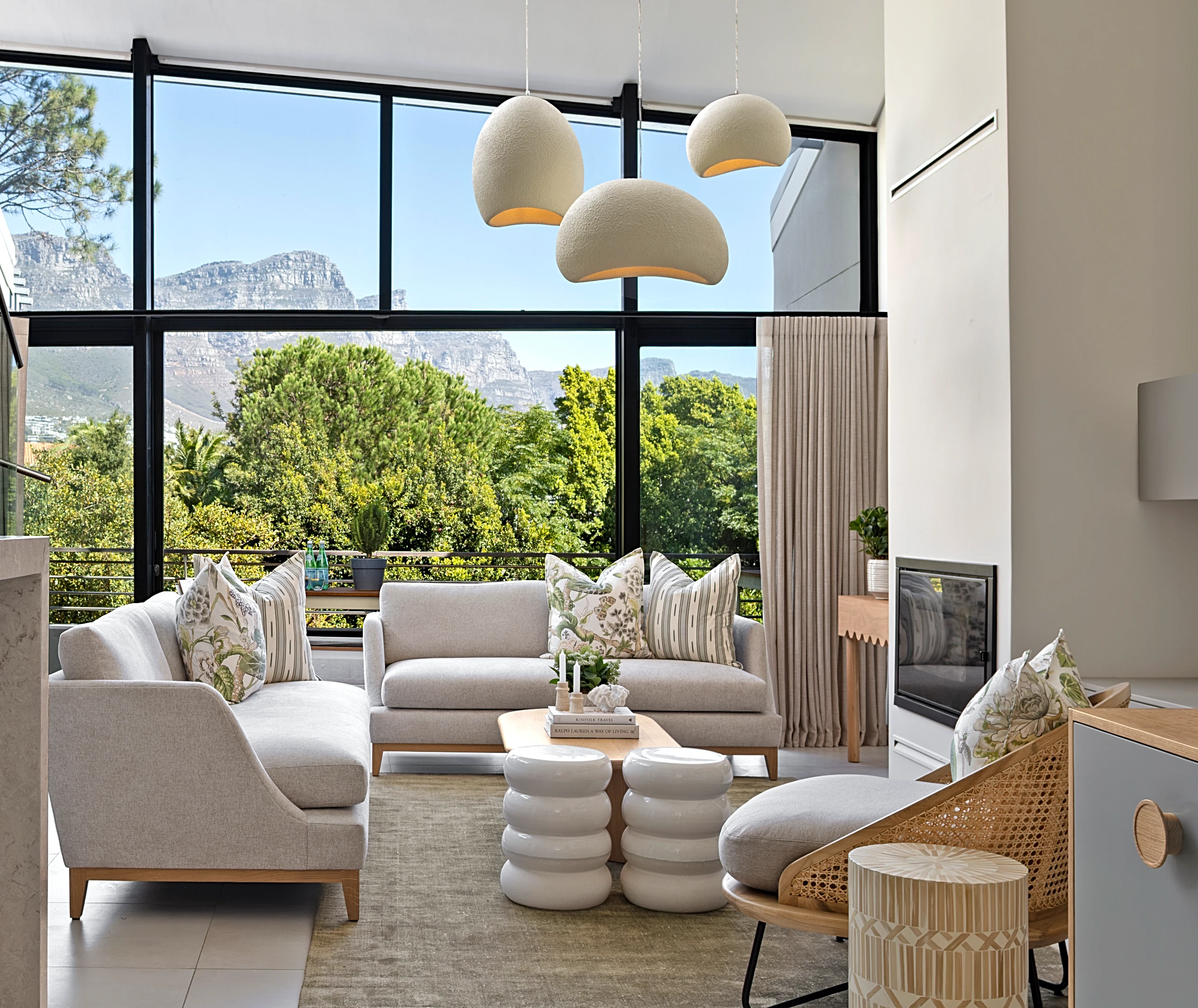 Living room at The Glen, Camps Bay: light sofas with botanical print cushions, three pendant lights and black-framed windows onto the trees and the mountain