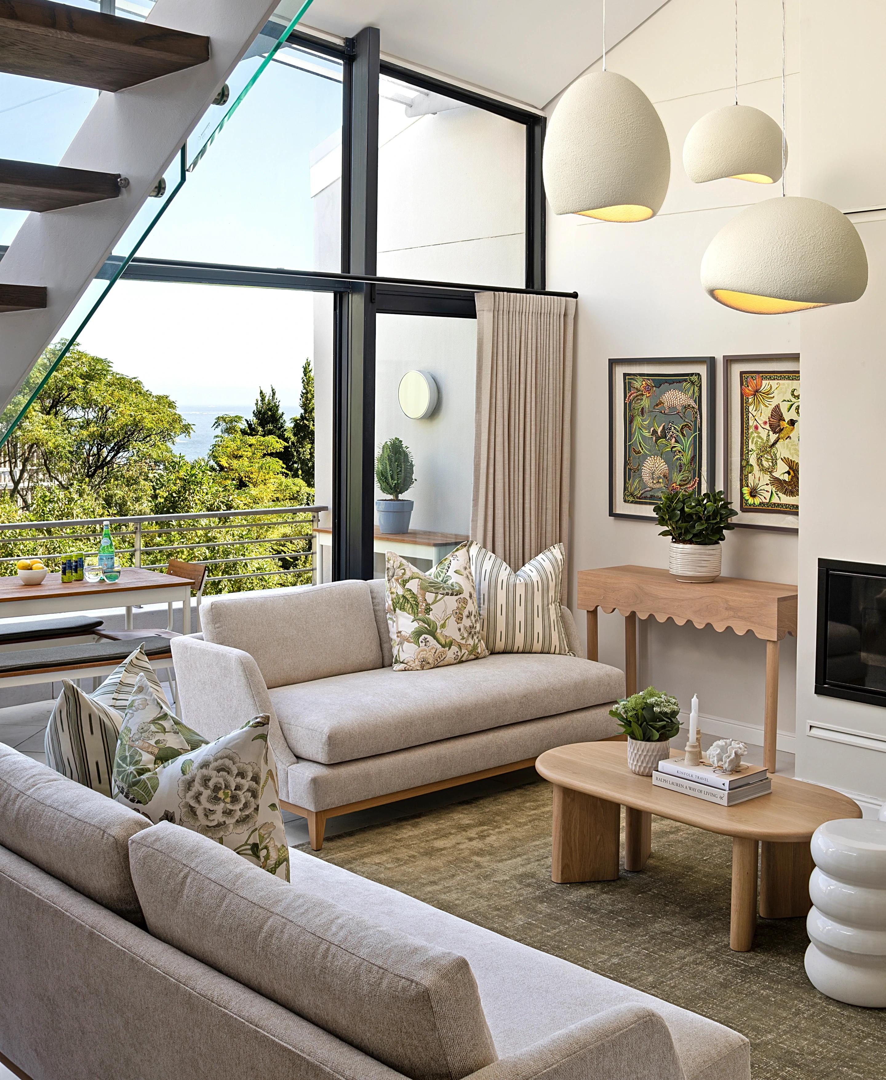 Living room at The Glen, Camps Bay: a light sofa with botanical print cushions, an oak coffee table and pendant lights below black-framed windows