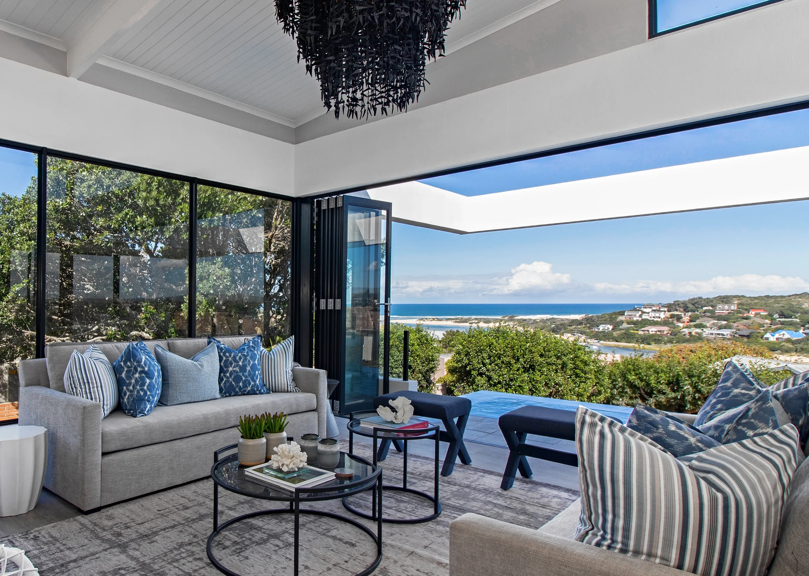 Living room at Kenton-on-Sea: a grey sofa with blue cushions, black nesting tables and a beaded chandelier, with folding doors open to the pool and the sea