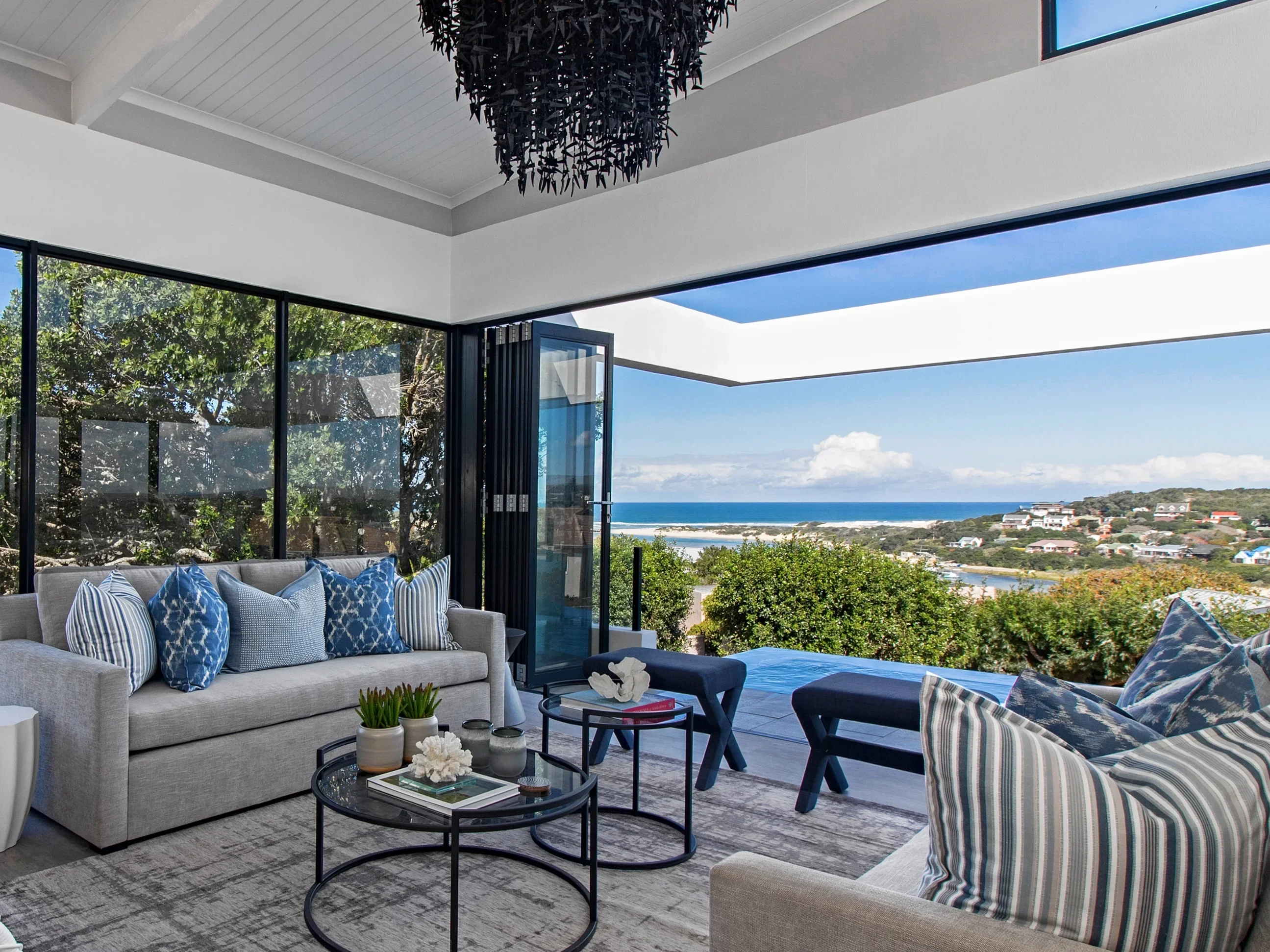Living room at Kenton-on-Sea: a grey sofa with blue cushions, black nesting tables and a beaded chandelier, with folding doors open to the pool and the sea