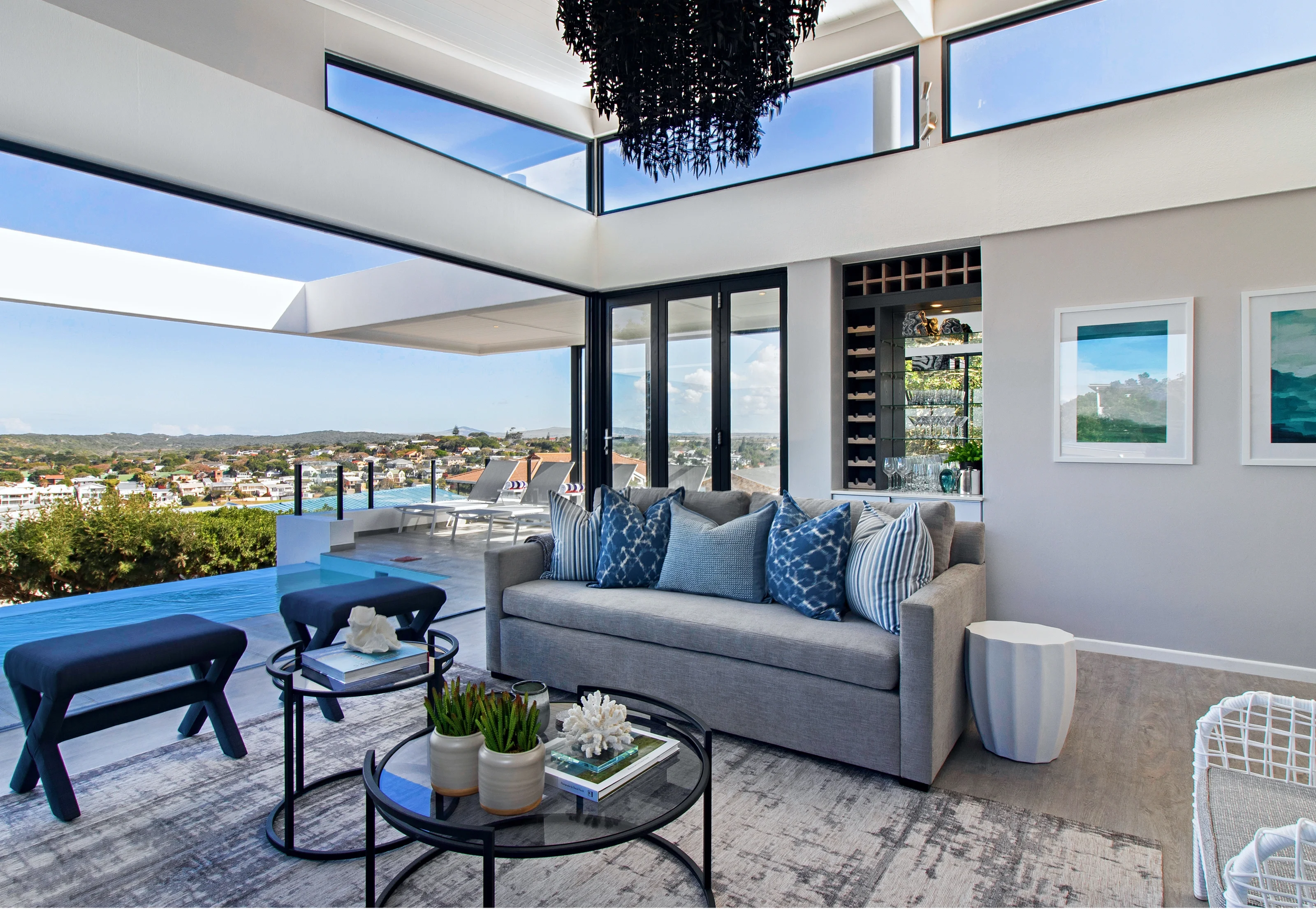 Covered terrace at Kenton-on-Sea, Eastern Cape: a grey sofa with blue cushions, black nesting tables and a view over the pool to the sea