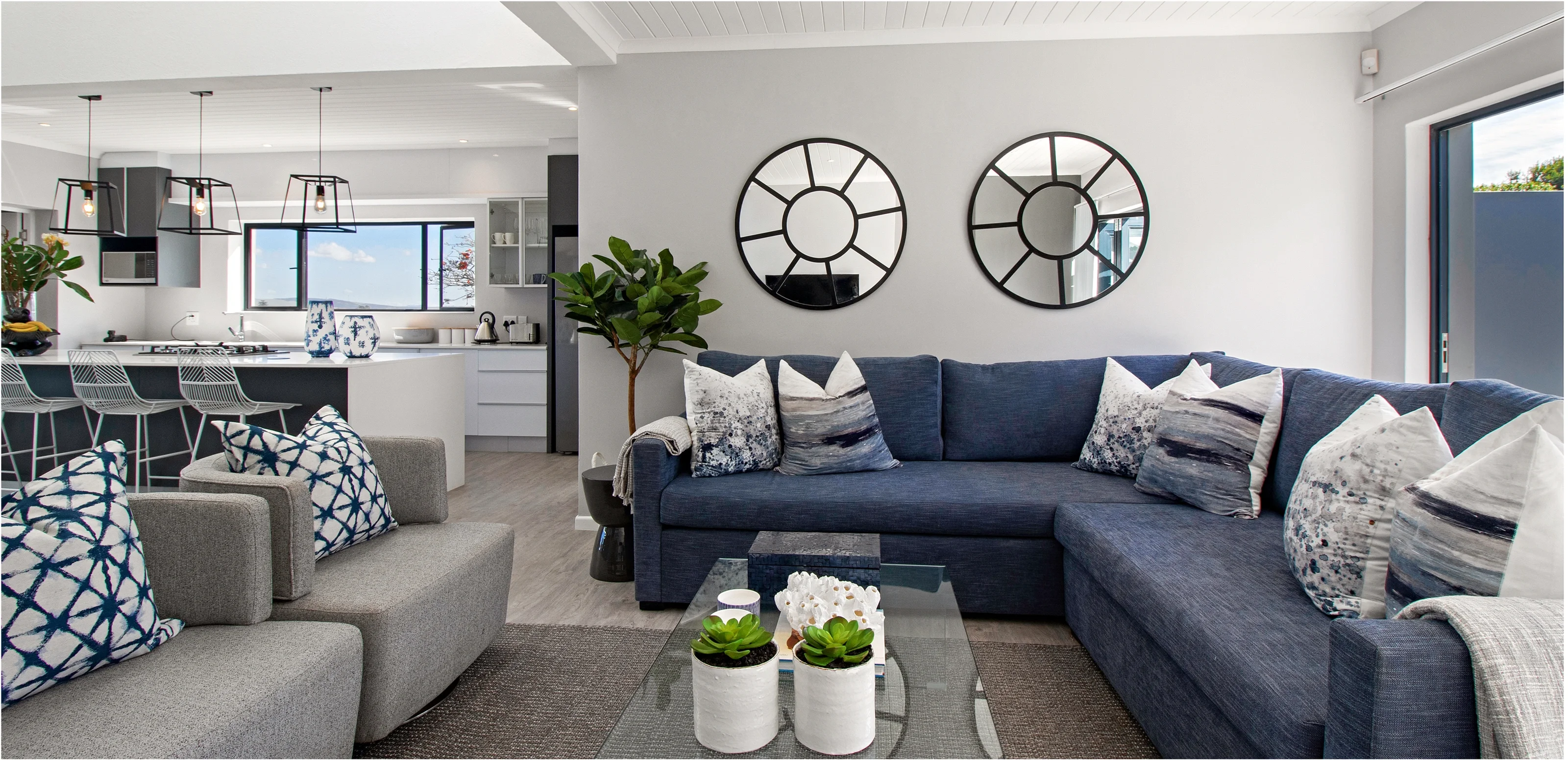 Living room at Kenton-on-Sea: a navy L-shaped sofa with patterned cushions, two round mirrors and the open kitchen beyond