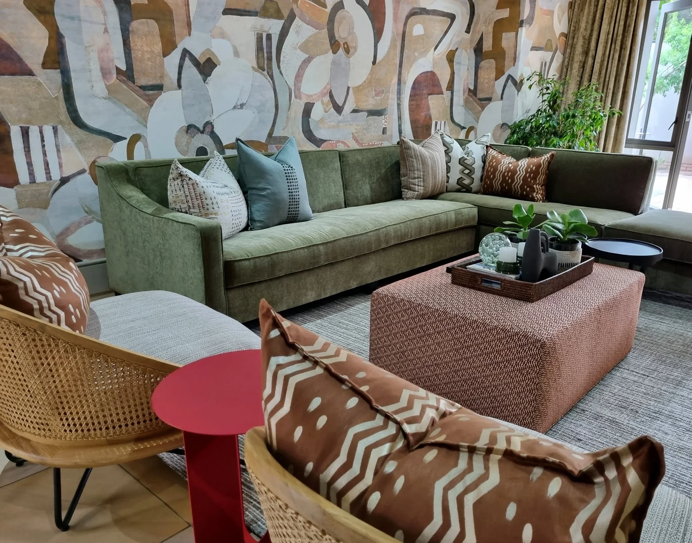 Living room with a large abstract mural, a green velvet sofa, a terracotta ottoman and rattan chairs